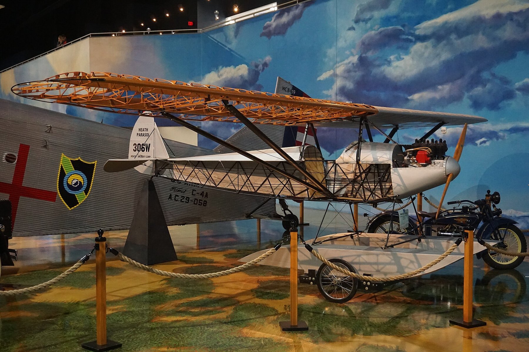 Figure 9 — A Heath Parasol displayed partly uncovered at the Air Zoo in Kalamazoo, Michigan, with its wooden wing ribs, steel-tube fuselage and float exposed and a Henderson motorcycle beside it. The tail is …
